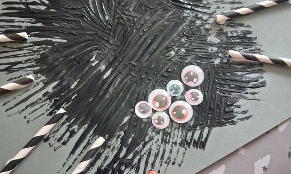 A gray paper with black paint spread like a spider, googly eyes, and paper straw spider legs attached with glue, plus a plastic fork. The background features decorative papers. @GLUEDTOMYCRAFTS is written at the bottom.