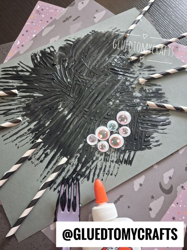 A gray paper with black paint spread like a spider, googly eyes, and paper straw spider legs attached with glue, plus a plastic fork. The background features decorative papers. @GLUEDTOMYCRAFTS is written at the bottom.