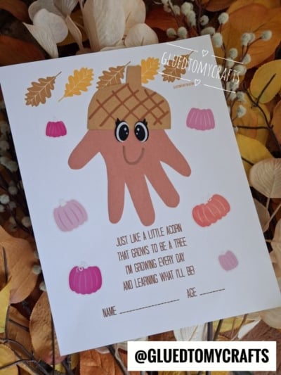 A handprint acorn craft on paper with cartoon eyes, surrounded by leaves and pumpkins. The text below features a sweet handprint acorn poem about growing like an acorn. There’s space for name and age at the bottom, all set against an autumn leaves background.
