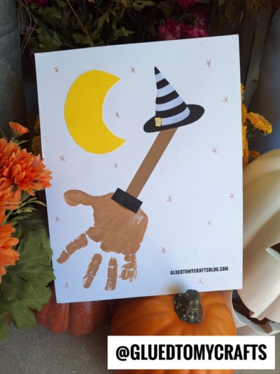 A Halloween-themed room on the broom craft features a handprint witch with a long arm acting as a handprint broom, black hat, crescent moon, orange flowers, pumpkin, and white jack-o-lantern. The text @GLUEDTOMYCRAFTS appears at the bottom.