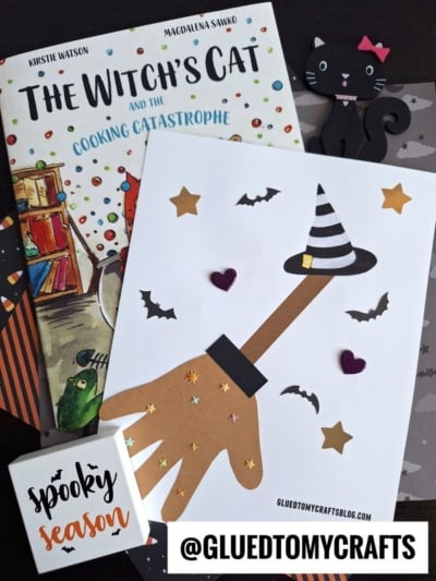 A childrens craft scene featuring The Witch’s Cat and the Cooking Catastrophe book, a handprint witch craft with hat and stars, a black cat decoration, a spooky season sign, plus a fun handprint broom for an extra touch of room on the broom craft magic!.