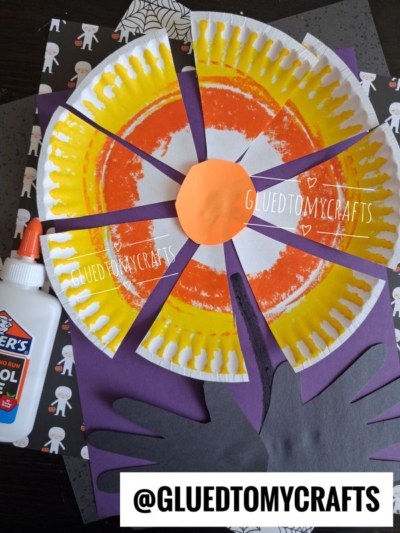 A colorful paper plate candy corn flower craft with orange and yellow paint, cut into sections, on purple and black paper, featuring a hand cutout, glue bottle, and a Halloween-themed background. @GLUEDTOMYCRAFTS watermark is present.