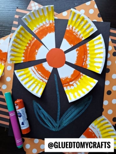 Colorful paper plate candy corn flower craft made from painted paper plates cut into petals, arranged on black paper with a drawn blue stem and leaves. Polka-dot orange and white background paper, markers, and glue stick are also visible.