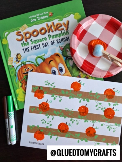 A children’s book titled “Spookley the Square Pumpkin,” a paper plate with orange paint and a pom-pom glued to a clothespin, a pom pom painted pumpkin patch craft with painted pumpkins on brown lines, and two markers are on a black surface.