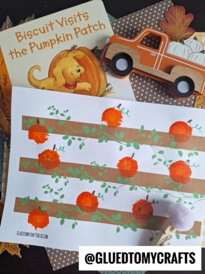A children’s book titled Biscuit Visits the Pumpkin Patch, a wooden toy truck with a pumpkin, and a pom pom painted pumpkin patch craft paper with orange stamps are arranged together on a table.