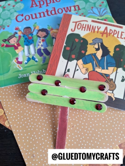 Two children’s books about apples are displayed alongside a creative popsicle stick tree craft, featuring red gems as apples on top, all arranged neatly on decorative paper. The text @GLUEDTOMYCRAFTS is at the bottom.