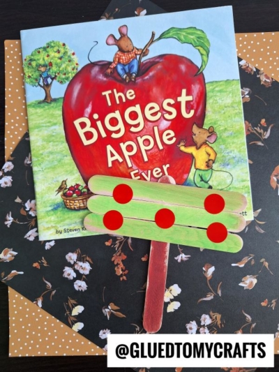 A childrens book titled The Biggest Apple Ever lies on a floral-patterned background. In front is a popsicle stick tree apple craft decorated with green sticks and red dot stickers. @GLUETOMYCRAFTS is written at the bottom.