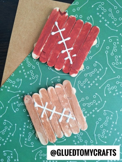 Two small popsicle stick footballs, decorated with brown and red paint and white laces, are displayed on green paper with play diagrams. The Instagram handle @GLUEDTOMYCRAFTS appears at the bottom.