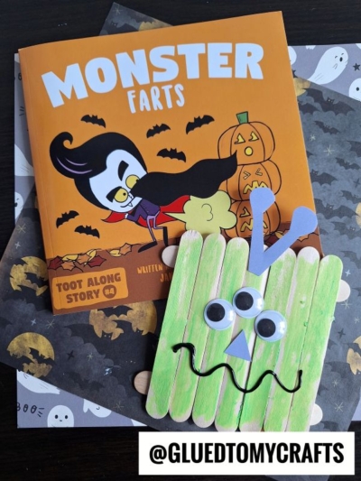 A childrens book titled Monster Farts with Halloween-themed illustrations sits next to a green popsicle stick monster craft with googly eyes and a zigzag mouth. The username @gluedtomycrafts is displayed at the bottom. Perfect for craft stick monsters fun!.