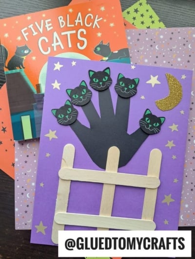 A Halloween craft features a black paper handprint with four black cat faces, a popsicle stick fence, and glittery moon and stars on purple paper. A Five Little Pumpkins book and patterned papers are in the background.