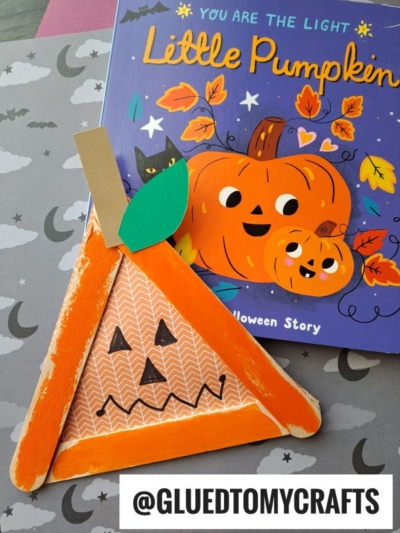 A children’s Halloween book with illustrated pumpkins sits beside a popsicle stick jack-o-lantern craft made from orange-painted sticks, patterned paper, a green leaf, and a cardboard stem. The username @gluedtomycrafts is shown at the bottom.