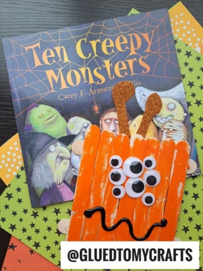 A Halloween book titled Ten Creepy Monsters is beside an orange craft stick monster, complete with googly eyes and glittery antennae, all placed on decorative paper. @GLUEDTOMYCRAFTS text is at the bottom.