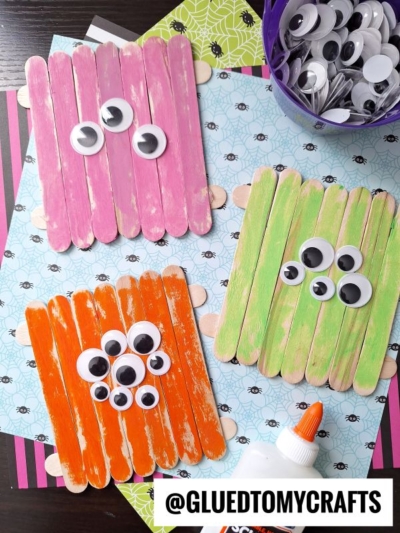 Three popsicle stick monsters made from colored sticks (pink, orange, green) with various sizes of googly eyes are displayed on patterned paper, alongside a glue bottle and a container of googly eyes.