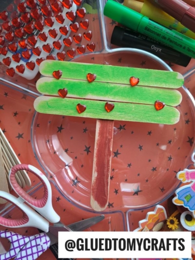 Craft supplies surround a popsicle stick tree, shaped like a tree and made from popsicle sticks colored green and brown, then decorated with red heart gems. Scissors, markers, and other craft items are also visible.