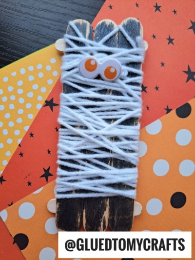 A popsicle stick mummy made from craft sticks painted black, wrapped with white yarn, and decorated with googly eyes. This fun craft is displayed on orange and yellow paper with playful polka dots and star patterns.