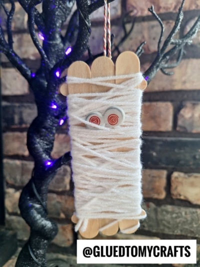 A popsicle stick mummy ornament made from craft sticks, googly eyes, and white yarn hangs in front of a black tree with purple lights. @GLUEDTOMYCRAFTS is written at the bottom.
