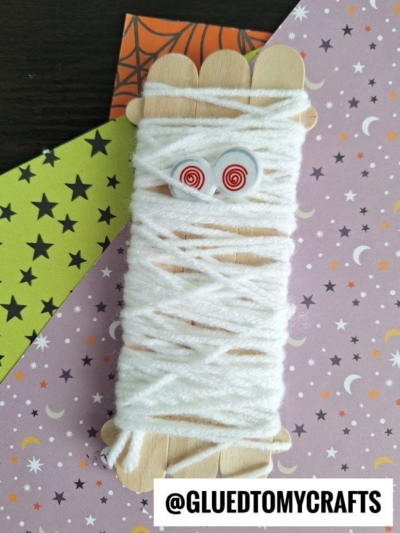 A popsicle stick mummy wrapped in white yarn with googly eyes rests on star- and moon-patterned papers. The text @GLUEDTOMYCRAFTS is at the bottom.