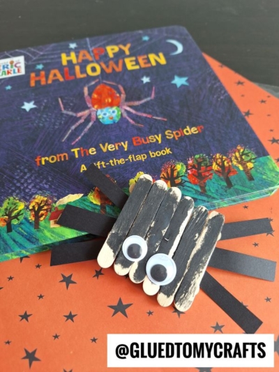 A Halloween-themed book and a popsicle stick spider craft made from black-painted sticks, googly eyes, and black paper legs are displayed on a star-patterned orange background. Text reads @GLUEDTOMYCRAFTS.
