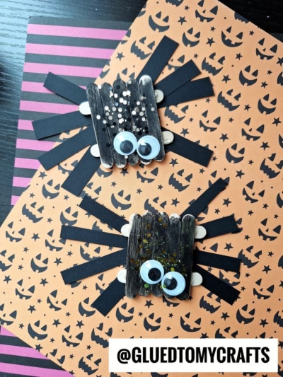 Two craft stick spider creations made from black popsicle sticks, googly eyes, and sparkles are displayed on orange paper with jack-o-lanterns and black striped purple paper. @GLUEDTOMYCRAFTS is written at the bottom.