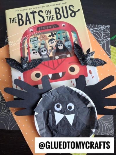 A children’s book titled The Bats on the Bus is shown with two glittery bat crafts and a black paper plate handprint bat, as well as a unique black bat made from paper and a bowl. The @GLUEDTOMYCRAFTS handle appears at the bottom.