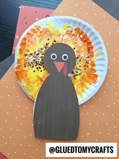 A colorful turkey craft made with a brown paper body, googly eyes, and a paper plate decorated with painted yellow and orange feathers and glitter, placed on top of patterned orange paper. This playful paint splat turkey brings vibrant charm to any fall display.