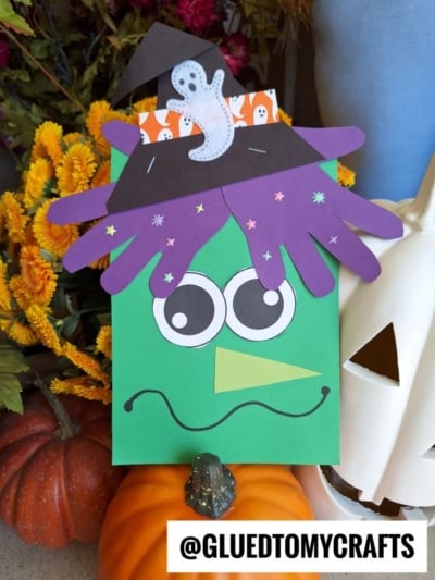 A green paper Frankenstein with googly eyes, purple hair, a witch hat, and a ghost is featured, surrounded by pumpkins and yellow flowers. Perfect as a handprint witch or paper witch keepsake craft for Halloween fun! @GLUEDTOMYCRAFTS is written at the bottom.