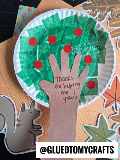 A paper plate apple tree with green tissue squares and red dots resembles a charming tree. A brown paper hand with Thanks for helping me grow! is attached. Fall-themed papers and @GLUEDTOMYCRAFTS are in the background.