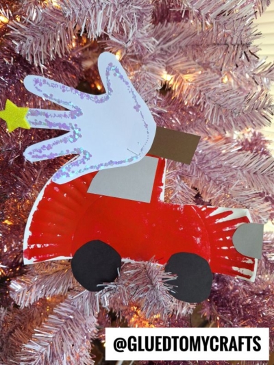 A handmade Christmas ornament of a red christmas car with black wheels, carrying a handprint cutout and a yellow star, hangs on a pink Christmas tree. The text @GLUEDTOMYCRAFTS appears at the bottom.