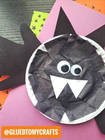 A paper plate handprint bat craft, made from a paper plate covered in black tissue with white paper fangs, googly eyes, and black paper wings and ears shaped like handprints, is displayed on colored paper.