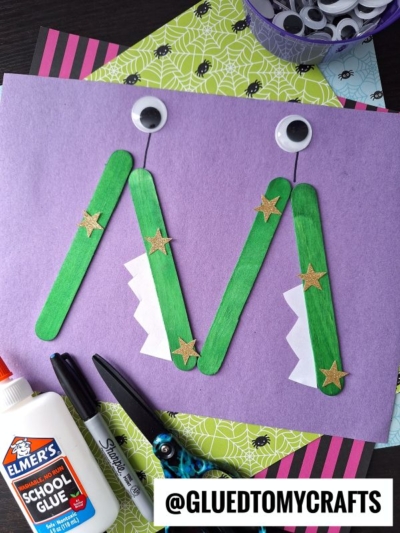 A letter M made of green craft sticks, decorated with gold stars, googly eyes, jagged white paper teeth—perfect for a fun “M is for Monster” craft—using school glue, markers, and scissors on a purple paper background. Instagram handle: @GLUEDTOMYCRAFTS.