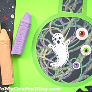 A green paper cutout shaped like a chalk art beaker is decorated with colored chalk squiggles, googly eyes, a white ghost, and a black bat.