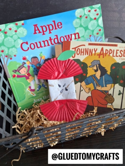 A black basket holds two children’s books, “Apple Countdown” and “Johnny Appleseed,” plus a toilet paper roll apple craft made from red cupcake liners and shredded brown filler. @GLUEDTOMYCRAFTS is on the bottom.