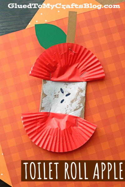 A craft tube apple made from a toilet paper roll, red cupcake liners, green paper leaf, and brown stem sits on an orange plaid background with the text TOILET ROLL APPLE.