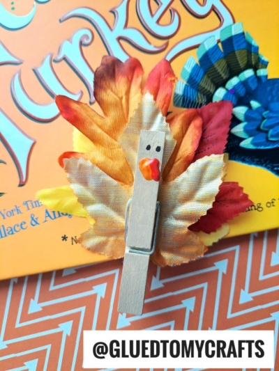 A leaf turkeys craft made from a clothespin, artificial autumn leaves for feathers, and googly eyes, displayed on an orange background with "Turkey" text and the handle @gluedtomycrafts.