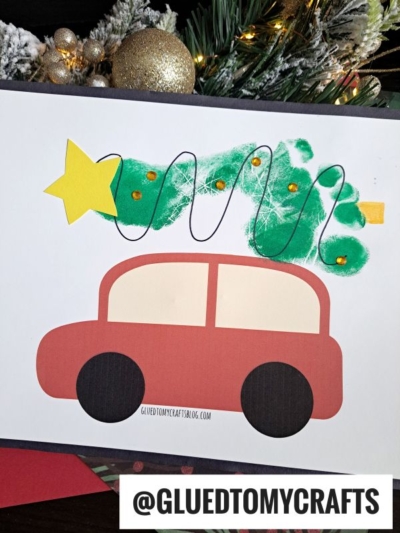 A children's craft featuring a red car with a green footprint Christmas tree on its roof, decorated with gold sequins, a yellow star, and black string, with holiday greenery in the background. @GLUEDTOMYCRAFTS is written at the bottom.