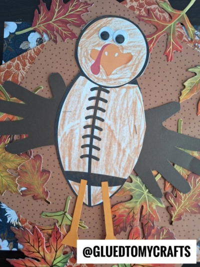A handmade football turkey craft made from colored paper, with handprints for wings, googly eyes, and a crayon-colored body, surrounded by autumn leaves. The text @GLUEDTOMYCRAFTS appears at the bottom.