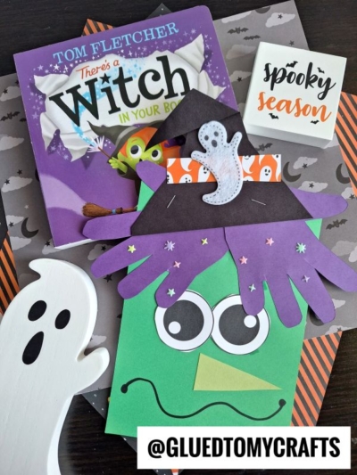 A Halloween-themed craft display with a book titled There's a Witch in Your Book, a handprint paper witch craft, a green monster face craft, wooden ghost shapes, and a Spooky Season sign on patterned paper.