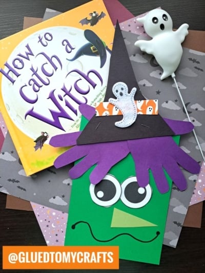A green handprint paper witch with big eyes, a purple hat, and a wavy mouth sits beside the book “How to Catch a Witch” and a white ghost decoration on dark, themed paper backgrounds.