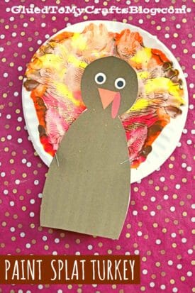 A colorful turkey craft made with a paper plate painted in red, orange, and yellow to mimic feathers, plus a brown paper body with googly eyes and beak, sits on a pink polka-dotted background. Text reads PAINT SPLAT TURKEY.