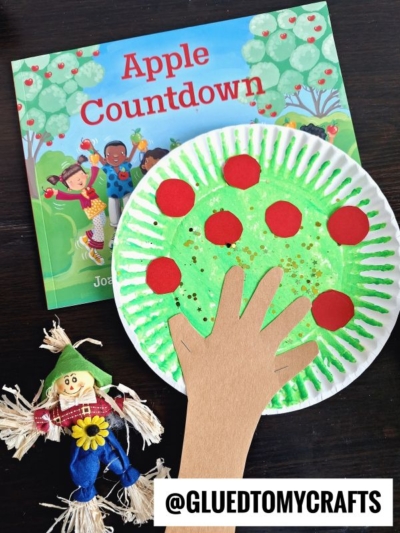 A children’s craft features a paper plate apple tree painted green with red dots, a paper hand cutout attached, an Apple Countdown book, and a small scarecrow doll; @GLUEDTOMYCRAFTS text is at the bottom.