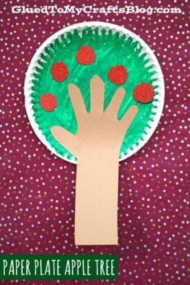 A paper plate apple tree painted green with red circles for apples forms the top, while a hand-shaped brown paper cutout serves as the trunk, all set against a red polka dot background.