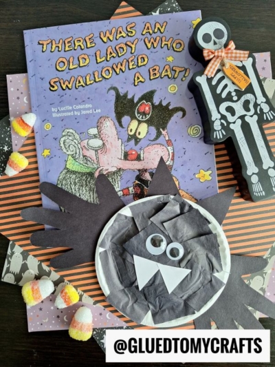 A Halloween-themed craft scene with the book There Was an Old Lady Who Swallowed a Bat!, a paper plate handprint bat craft, a skeleton figure, candy corns, and festive paper, all displayed on a dark surface.