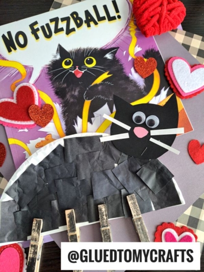 A children’s craft scene featuring the book “No Fuzzball!” with a black cat on the cover, a handmade clothespin black cat craft with googly eyes, and heart decorations. The handle @GLUEDTOMYCRAFTS is at the bottom.