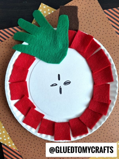 A paper plate felt apple craft featuring red felt squares around the edge, a green felt handprint for the leaf, a brown felt stem, and drawn black seeds in the center—perfect for creative fun!.