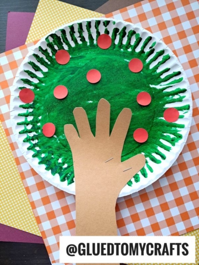 A paper plate apple tree painted green with red circles, resembling an apple tree. A brown paper cut-out hand forms the trunk, and the plate sits on orange and yellow plaid papers. @GLUEDTOMYCRAFTS is at the bottom.