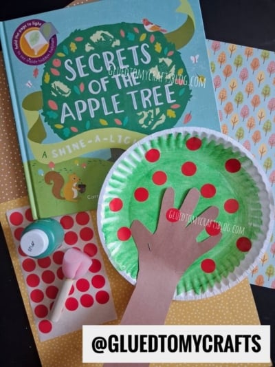 A children’s book, Secrets of the Apple Tree, is shown alongside a delightful paper plate apple tree craft, featuring red sticker dots, a brown paper hand cutout, paint, and colorful paper—all arranged on a tabletop. @GLUEDTOMYCRAFTS is labeled.