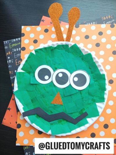 A Paper Plate Monster craft featuring a green, three-eyed monster with orange glittery antennae, a triangular nose, and a wavy mouth, displayed on orange and black Halloween-themed paper. @GLUEDTOMYCRAFTS is written at the bottom.