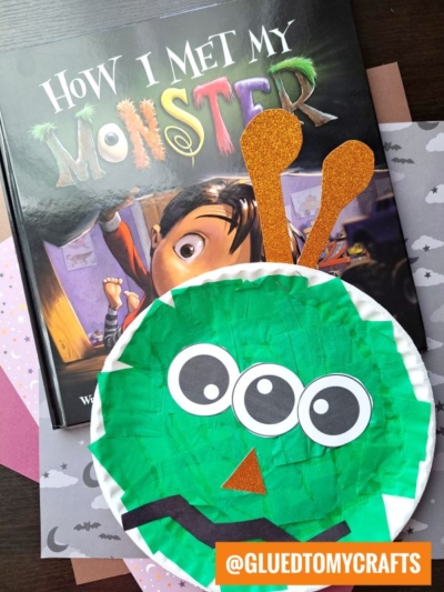 A children’s book titled How I Met My Monster sits next to a colorful Paper Plate Monster craft featuring big eyes, orange antennae, and a triangle nose. The playful background is decorated with star and cloud paper.