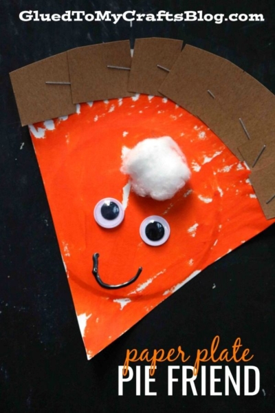 A paper plate pumpkin pie craft resembling a pie slice with orange paint, googly eyes, a cotton ball whipped cream, cardboard crust, and a drawn smile. Text reads: paper plate PIE FRIEND.
