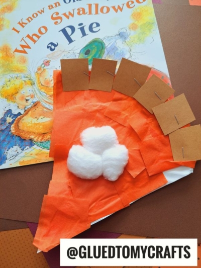A children’s book titled “I Know an Old Lady Who Swallowed a Pie” lies beneath a pumpkin pie paper plate craft, featuring orange tissue for filling, brown squares as crust, and cotton balls as whipped cream.